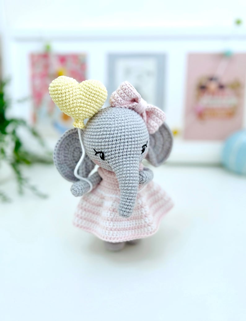Peut inclure: Un &eacute;l&eacute;phant en crochet gris portant une robe ray&eacute;e rose et blanche et un n&oelig;ud rose. L'&eacute;l&eacute;phant tient un ballon jaune en forme de c&oelig;ur.
