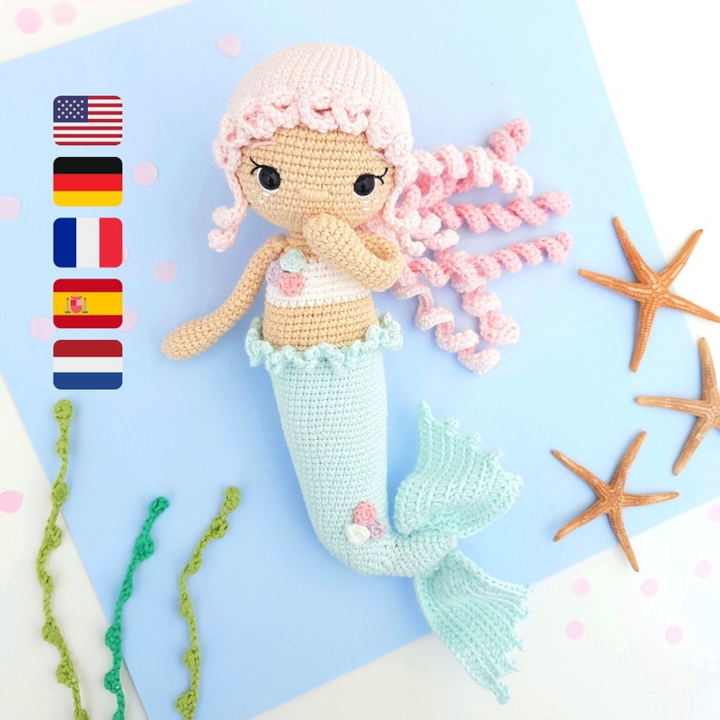 May include: A crocheted mermaid doll with pink hair, a light blue tail, and a pink and white seashell top. The doll is sitting on a blue background with starfish.