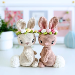 Peut inclure: Deux lapins en crochet portant des couronnes de fleurs. Le lapin à gauche est beige et le lapin à droite est brun clair. Les deux lapins ont des queues blanches et duveteuses.