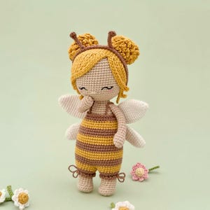 Peut inclure: Une poup&eacute;e crochet&eacute;e portant une tenue sur le th&egrave;me de l'abeille avec des rayures horizontales jaunes et brunes, des ailes blanches et un bonnet jaune avec deux antennes. La poup&eacute;e a des yeux ferm&eacute;s brod&eacute;s, des joues roses et des fleurs.