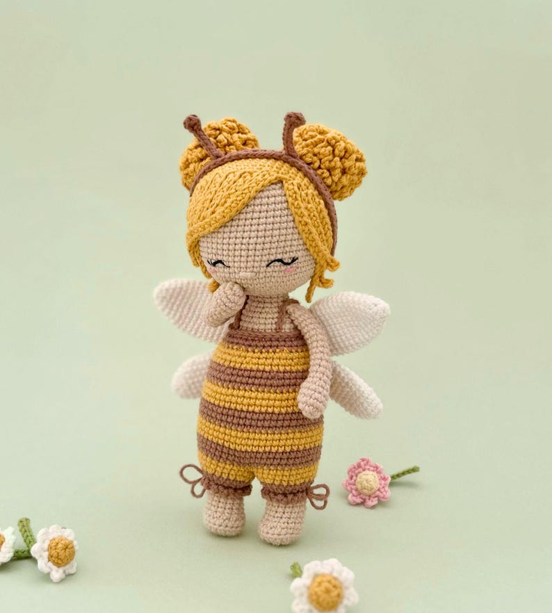 Peut inclure: Une poup&eacute;e crochet&eacute;e portant une tenue sur le th&egrave;me de l'abeille avec des rayures horizontales jaunes et brunes, des ailes blanches et un bonnet jaune avec deux antennes. La poup&eacute;e a des yeux ferm&eacute;s brod&eacute;s, des joues roses et des fleurs.