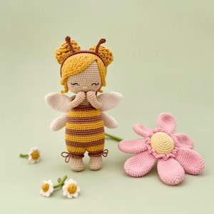 Peut inclure: Une poup&eacute;e crochet&eacute;e d&eacute;guis&eacute;e en abeille avec des rayures jaunes et brunes, une peau claire et de petites ailes blanches. La poup&eacute;e a des cheveux jaunes, des antennes et est plac&eacute;e &agrave; c&ocirc;t&eacute; d'une grande fleur crochet&eacute;e rose et trois petites marguerites.