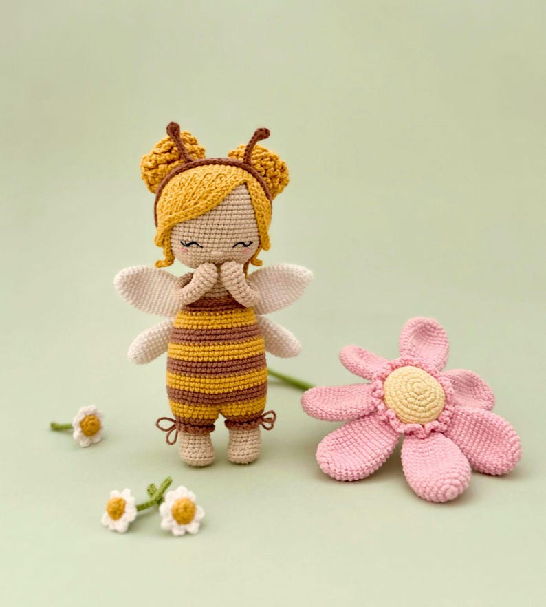 Peut inclure: Une poup&eacute;e crochet&eacute;e d&eacute;guis&eacute;e en abeille avec des rayures jaunes et brunes, une peau claire et de petites ailes blanches. La poup&eacute;e a des cheveux jaunes, des antennes et est plac&eacute;e &agrave; c&ocirc;t&eacute; d'une grande fleur crochet&eacute;e rose et trois petites marguerites.