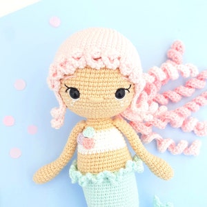 May include: A crocheted mermaid doll with pink hair, a light blue tail, and a white and pink top. The doll has black eyes and a crocheted face.