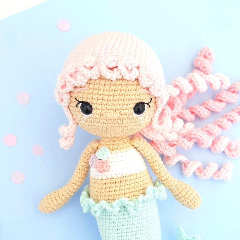 May include: A crocheted mermaid doll with pink hair, a light blue tail, and a white and pink top. The doll has black eyes and a crocheted face.