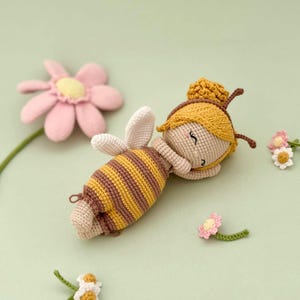 Peut inclure: Une poup&eacute;e crochet&eacute;e en forme d'abeille, avec un corps ray&eacute; jaune et marron, des ailes blanches et un bonnet jaune &agrave; antennes marron. La poup&eacute;e a un visage beige aux yeux ferm&eacute;s et est entour&eacute;e de plusieurs petites fleurs crochet&eacute;es roses et blanches.