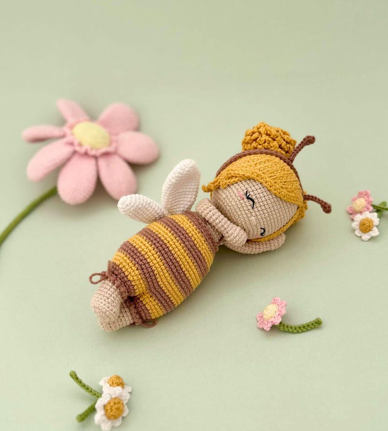 Peut inclure: Une poup&eacute;e crochet&eacute;e en forme d'abeille, avec un corps ray&eacute; jaune et marron, des ailes blanches et un bonnet jaune &agrave; antennes marron. La poup&eacute;e a un visage beige aux yeux ferm&eacute;s et est entour&eacute;e de plusieurs petites fleurs crochet&eacute;es roses et blanches.