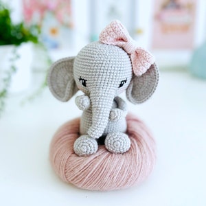 Peut inclure: Un &eacute;l&eacute;phant en crochet gris avec des oreilles en laine rose et un n&oelig;ud rose. L'&eacute;l&eacute;phant est assis sur une boule de laine rose.