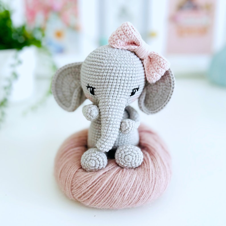 Peut inclure: Un &eacute;l&eacute;phant en crochet gris avec des oreilles en laine rose et un n&oelig;ud rose. L'&eacute;l&eacute;phant est assis sur une boule de laine rose.