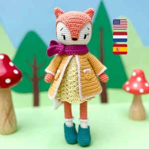 Può includere: Una bambola di volpe all'uncinetto che indossa un cappotto giallo, una sciarpa rosa e un vestito giallo con un bordo rosa. La bambola è in piedi su una superficie verde con due decorazioni a forma di fungo.