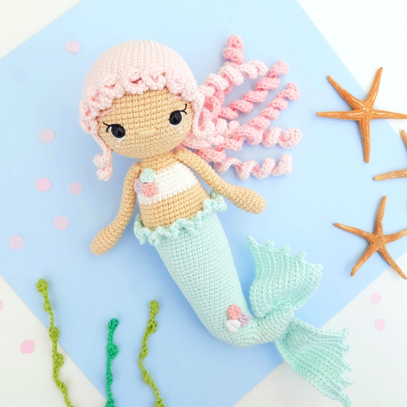 May include: A crocheted mermaid doll with pink hair, a light blue tail, and a white and pink top. The doll is on a light blue background with green seaweed and starfish.