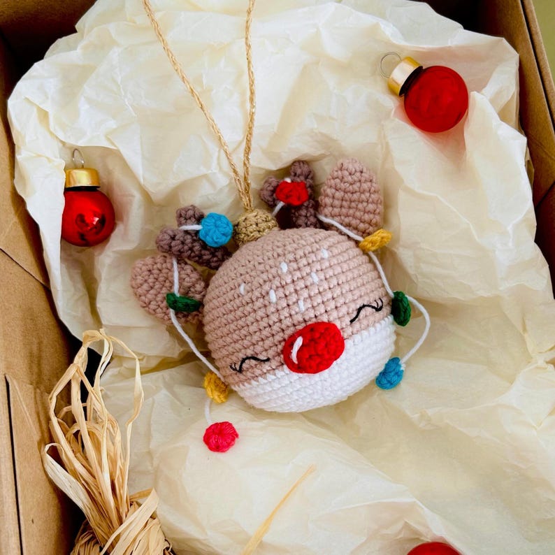 May include: A handmade crochet reindeer pendant necklace with a gold chain. The reindeer has a tan head, white face, red nose, and colorful lights. Two red Christmas ornaments are also in the image.