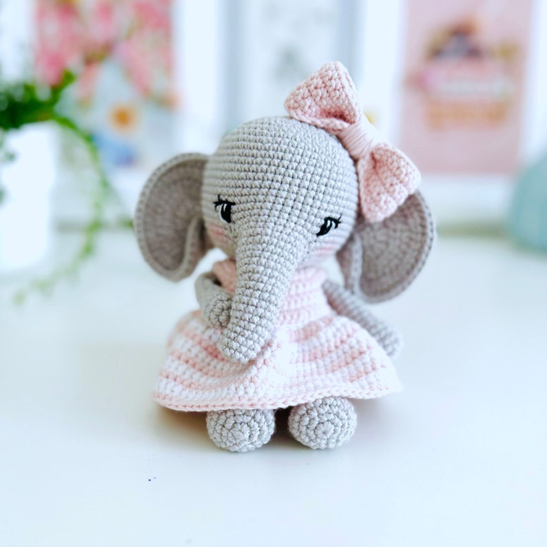 Peut inclure: Un &eacute;l&eacute;phant en crochet gris portant une robe ray&eacute;e rose et blanche avec un n&oelig;ud rose sur la t&ecirc;te.