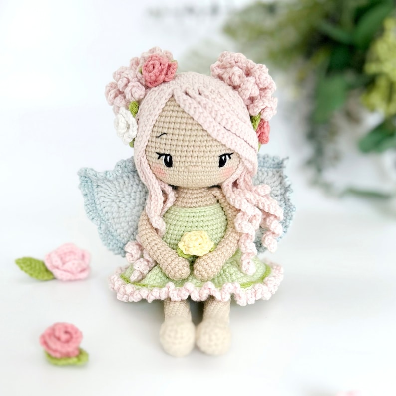 May include: A crocheted fairy doll with pink hair, a green dress, and blue wings. The doll is holding a yellow flower and is sitting on a white surface.