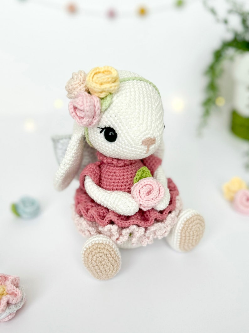 Puede incluir: Un conejito de crochet blanco con un vestido rosa con ribete de encaje blanco y una corona de flores. El conejito sostiene una flor rosa en sus patas.