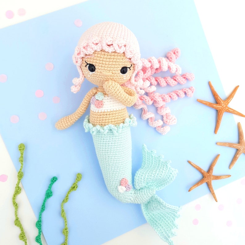 May include: A crocheted mermaid doll with pink hair, a light blue tail, and a seashell-decorated bodice. The doll is sitting on a light blue background with green seaweed and pink confetti.