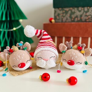 May include: Crocheted Christmas ornaments featuring Santa Claus and reindeer. The Santa ornament has a red and white striped hat. The reindeer have red noses and are adorned with miniature string lights. Red Christmas ball ornaments are scattered around.