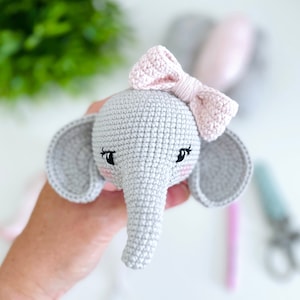Peut inclure: Un &eacute;l&eacute;phant en crochet gris avec des oreilles roses et un n&oelig;ud rose. L'&eacute;l&eacute;phant a des yeux brod&eacute;s noirs et des joues roses.
