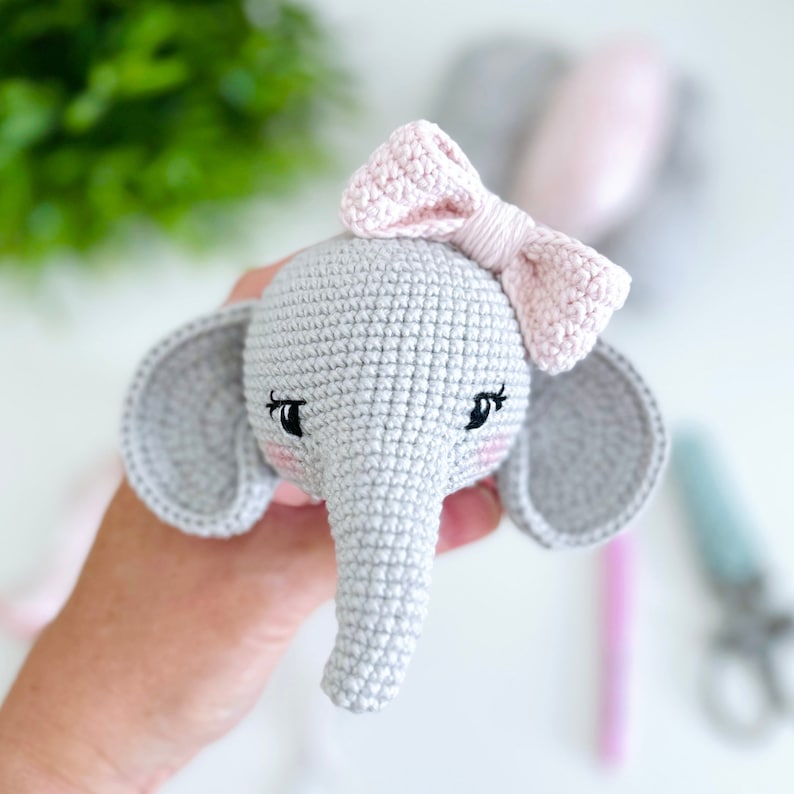 Peut inclure: Un &eacute;l&eacute;phant en crochet gris avec des oreilles roses et un n&oelig;ud rose. L'&eacute;l&eacute;phant a des yeux brod&eacute;s noirs et des joues roses.