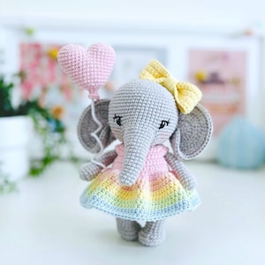 Peut inclure: Un &eacute;l&eacute;phant en crochet gris portant une robe rose et arc-en-ciel et un n&oelig;ud jaune. L'&eacute;l&eacute;phant tient un ballon rose en forme de c&oelig;ur.