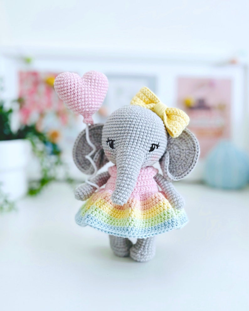 Peut inclure: Un &eacute;l&eacute;phant en crochet gris portant une robe rose et arc-en-ciel et un n&oelig;ud jaune. L'&eacute;l&eacute;phant tient un ballon rose en forme de c&oelig;ur.