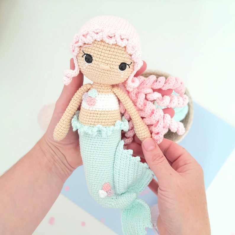 May include: A crocheted mermaid doll with pink hair, a light blue tail, and a white and pink top. The doll is holding a seashell in its hand.