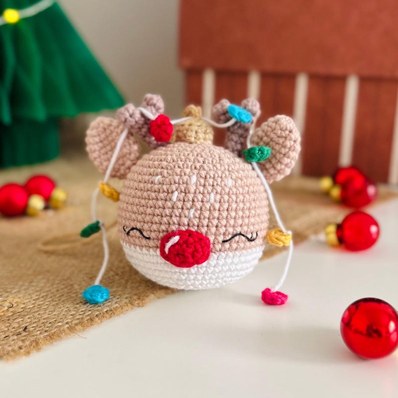 May include: A crocheted Christmas ornament shaped like a reindeer with a red nose and string lights. The reindeer is beige with white at the bottom. Red Christmas ornaments are scattered around the reindeer.