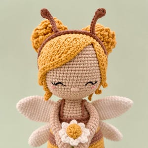 Peut inclure: Une poup&eacute;e crochet&eacute;e au design inspir&eacute; d'une abeille avec des v&ecirc;tements ray&eacute;s jaunes et marron, des ailes claires et un bandeau avec des antennes. La poup&eacute;e a la peau claire, les yeux ferm&eacute;s avec des cils, des joues roses et tient une petite fleur blanche.