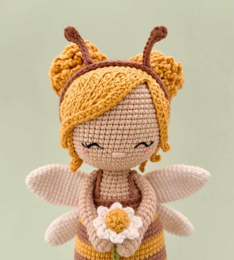 Peut inclure: Une poup&eacute;e crochet&eacute;e au design inspir&eacute; d'une abeille avec des v&ecirc;tements ray&eacute;s jaunes et marron, des ailes claires et un bandeau avec des antennes. La poup&eacute;e a la peau claire, les yeux ferm&eacute;s avec des cils, des joues roses et tient une petite fleur blanche.