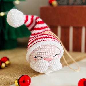 May include: A crocheted Santa Claus ornament with a white beard and a red and white striped hat. The ornament has a smiling face with closed eyes and a pink nose. A white pom-pom tops the hat. A jute string is attached for hanging. Christmas decor.