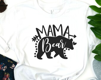 May include: White t-shirt with a black graphic that reads "MAMA Bear". The design features a bear silhouette with an arrow and decorative elements. The shirt is styled with sunglasses, floral pants, and gray sneakers.