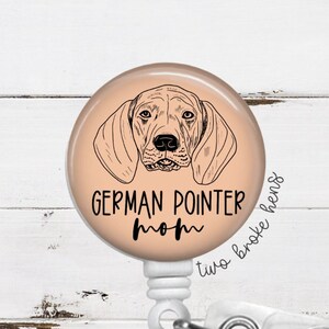 May include: A peach-colored badge holder with a black and white illustration of a German Shorthaired Pointer dog. The text "German Pointer Mom" is printed on the badge holder. The text "two broke hens" is printed on the side of the badge holder.