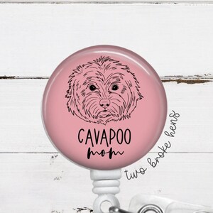 May include: Pink round badge holder with a black and white illustration of a Cavapoo dog. The text "Cavapoo Mom" is written below the dog. The badge holder is attached to a clear plastic retractable badge reel.