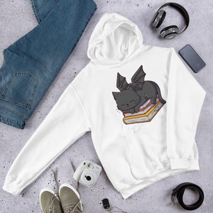 K&ouml;nnte beinhalten: Wei&szlig;er Kapuzenpullover mit einer schwarzen Katze mit Fl&uuml;geln, die auf einem Stapel B&uuml;cher schl&auml;ft. Die Katze hat einen niedlichen Cartoon-Stil.