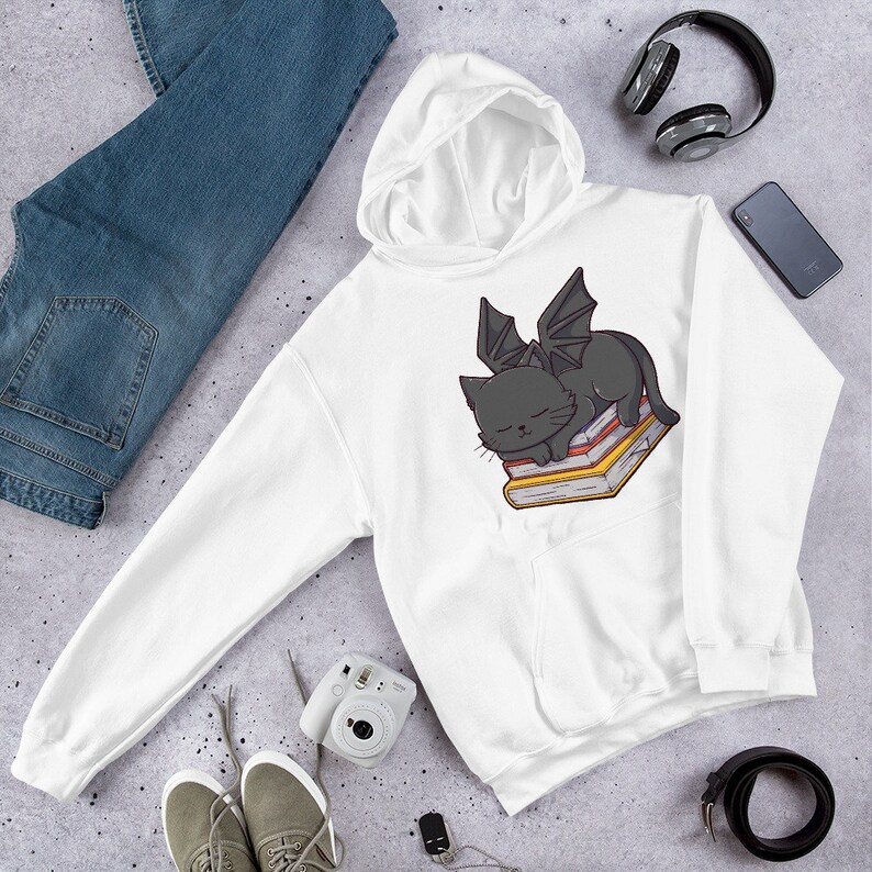 K&ouml;nnte beinhalten: Wei&szlig;er Kapuzenpullover mit einer schwarzen Katze mit Fl&uuml;geln, die auf einem Stapel B&uuml;cher schl&auml;ft. Die Katze hat einen niedlichen Cartoon-Stil.