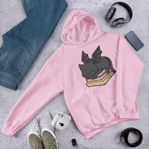 K&ouml;nnte beinhalten: Rosa Kapuzenpullover mit einer schwarzen Katze mit Fl&uuml;geln, die auf einem Stapel B&uuml;cher schl&auml;ft. Die Katze hat einen s&uuml;&szlig;en Gesichtsausdruck.