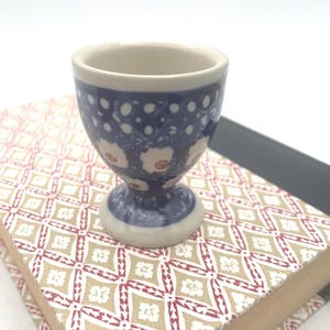 May include: A ceramic egg cup featuring a dark blue glaze with a white polka dot pattern on the upper rim and a repeating floral motif with orange centers on the body, set on a pedestal base with a smooth, off-white interior finish.