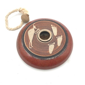 May include: A round, reddish-brown ceramic incense burner with a central hole and a decorative design. The design includes a figure playing a wind instrument and footprints. A beige rope with a small, round bead is attached.