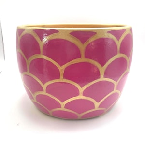May include: A round, pink ceramic planter with a gold, scale-like pattern. The interior and rim of the planter are a light gold color. The planter is a decorative item for indoor or outdoor use.