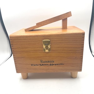 May include: A wooden Ronson Roto-Shine Magnetic shoe shine box with a hinged lid and a wooden footrest. The box is made of light brown wood and has a brass clasp. The text "Ronson Roto-Shine Magnetic" is printed on the front.