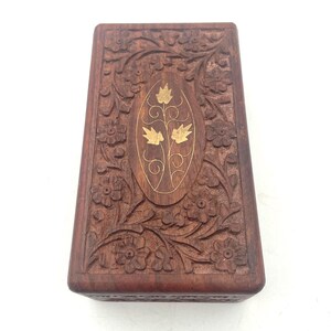 May include: A rectangular brown wooden box featuring intricate floral carvings across the lid. The center of the lid displays an oval inlay of smooth wood decorated with a gold-colored metal leaf motif. The box has a dark, natural wood grain finish.