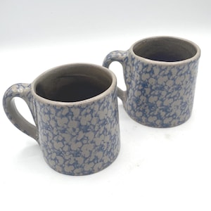 May include: Two ceramic mugs with a speckled blue and gray pattern. Each mug has a handle and a wide opening. The mugs are set against a white background.