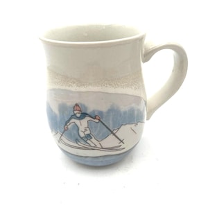 May include: A ceramic mug with a cream-colored glaze on the upper portion and a hand-painted illustration of a skier in a white suit, blue accents, and a red hat on a snowy mountain landscape on the lower portion, featuring a curved handle.