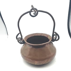 May include: A small, round, hammered copper pot featuring a dark, wrought iron swing handle. The handle is attached to the pot with decorative, curled iron loops on each side, and the pot has a wide, open mouth with a slightly flared rim.