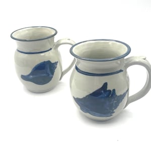 May include: Two ceramic mugs with a white base, blue rim, and a blue seashell design. Each mug has a curved handle and a blue stripe around the middle. The mugs are set against a white background.