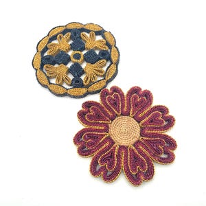 May include: Two decorative coasters. One is a circular design with gold and navy blue woven details. The other is a flower shape with burgundy and gold woven details and a tan center. Both are on a white background.