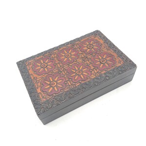 May include: A rectangular wooden box with a floral design. The box features a dark brown exterior and a top panel with a repeating pattern of red and orange flowers. The edges of the box are decorated with a carved floral pattern.