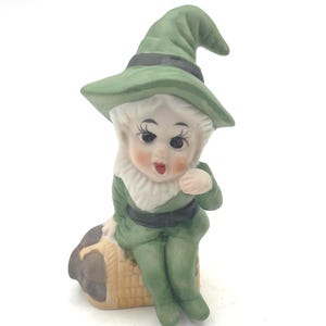 May include: A ceramic figurine of a seated elf wearing a green hat and outfit with a black belt. The elf has a white beard and rosy cheeks. It is sitting on a basket-like object. The figurine is on a white background.