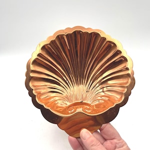 May include: A copper-colored, shell-shaped decorative dish. The dish has a scalloped edge and a detailed fan-like pattern radiating from the center. The dish is held by a hand against a white background.