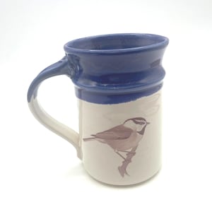 May include: A ceramic mug with a glossy dark blue rim and handle, featuring a cream-colored body decorated with a detailed brown illustration of a small bird perched on a branch. The mug has a cylindrical shape with a slightly flared top edge.